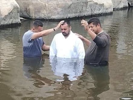 Una persona vestida de blanco de pie en un río mientras dos hombres la bautizan.