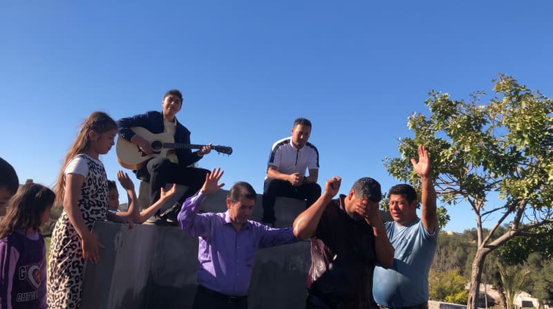 Personas con las manos levantadas y un hombre tocando la guitarra junto a una alberca de bautismo.