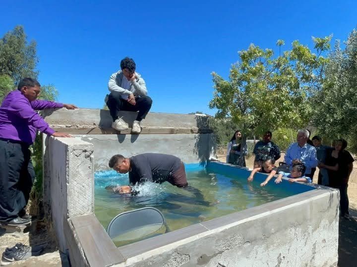 Una persona siendo bautizada en una alberca al aire libre mientras otros observan.