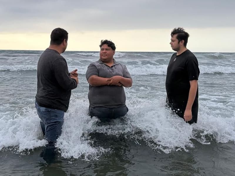 Un bautismo en el oleaje: tres hombres de pie entre las olas bajo un cielo nublado; el del centro, con los brazos cruzados.
