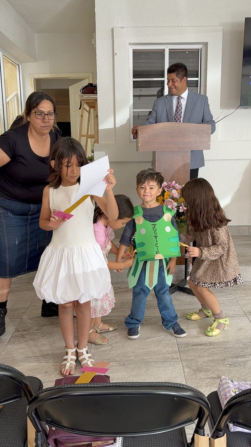 A teacher helping a girl with a paper while a small boy in a green paper armor stands beside her, with the pastor behind the pulpit.