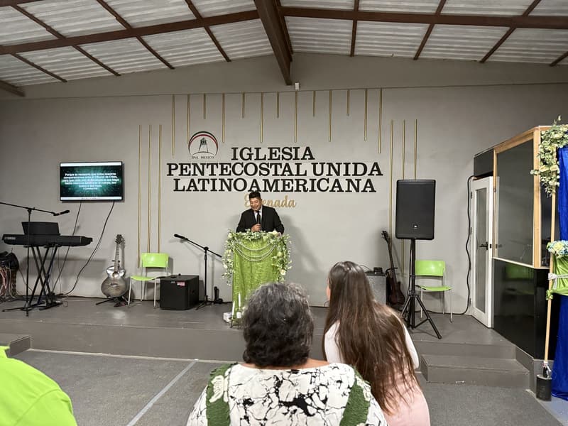 A service in the Ensenada church hall, with a man speaking at the pulpit under the church name.