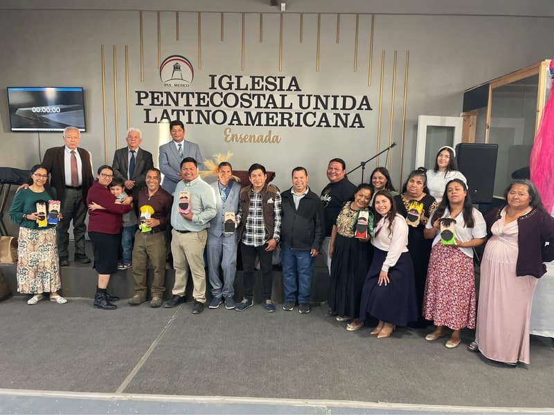 Church members of all ages standing together in front of the church name on the wall at the Ensenada church, several holding handmade crafts.