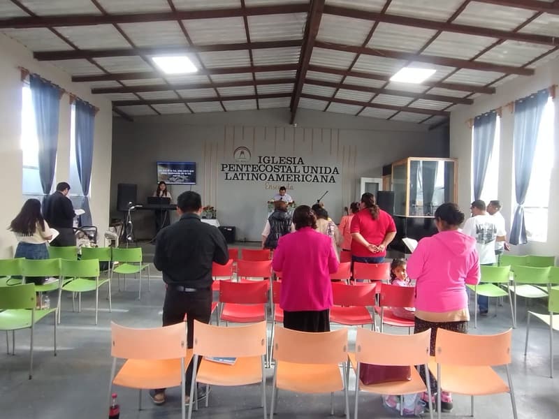 Worshipers standing in the Ensenada church hall, seen from behind, facing the stage and the church name.