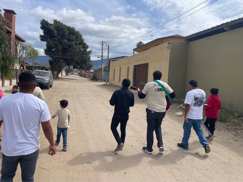 Members and children walking down a neighborhood dirt street, one carrying a guitar, seen from behind.
