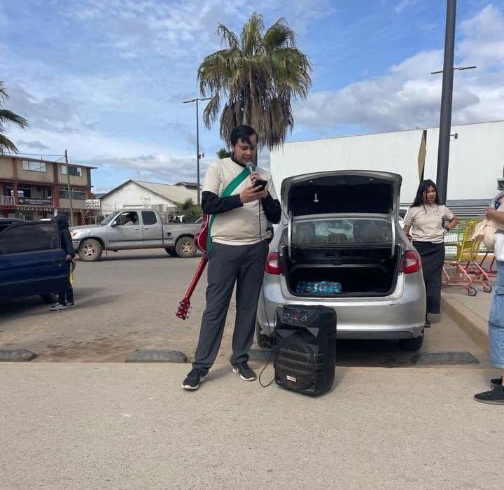 A young man with a guitar on his back stands by the open trunk of a car and a speaker in a parking lot, with a woman nearby.