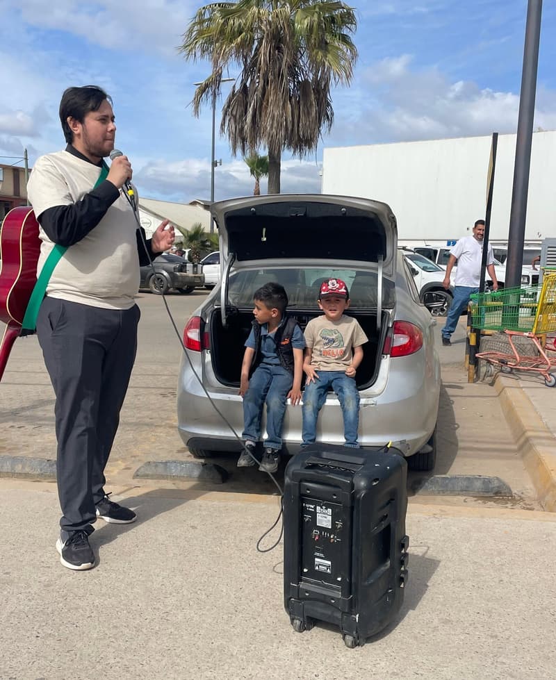 A young man preaching with a microphone in a parking lot, a guitar on his back, while two boys sit in the open trunk of a car beside a speaker.