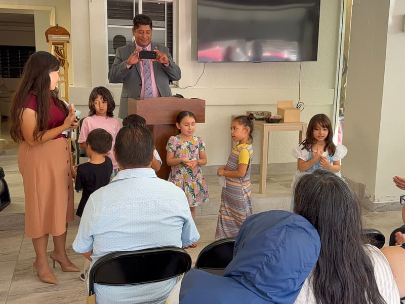 Children stand at the front of the Tijuana church with their teacher, holding papers as they present, while the pastor takes a photo and the congregation watches.