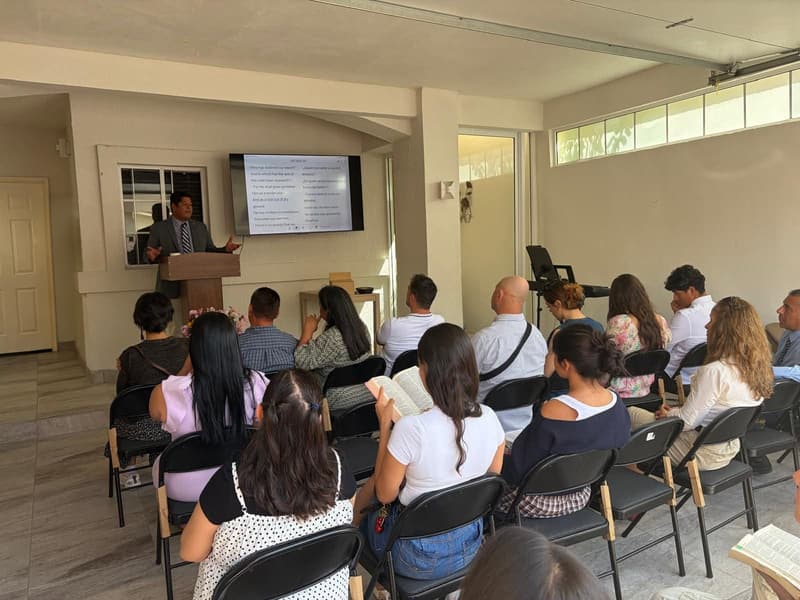 A service at IPUL Tijuana: a man preaches at the pulpit, with Bible verses in English and Spanish on the screen behind him, while the congregation listens, some with open Bibles.